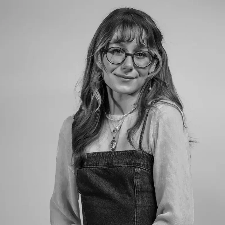 A young woman with glasses and wavy hair smiling, wearing layered jewelry and a denim dress over a long-sleeve shirt.