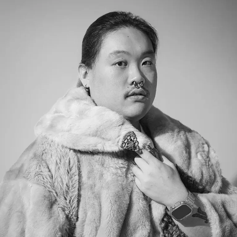 A person with piercings and a tattoo, wearing a fur coat, looking serious at the camera.