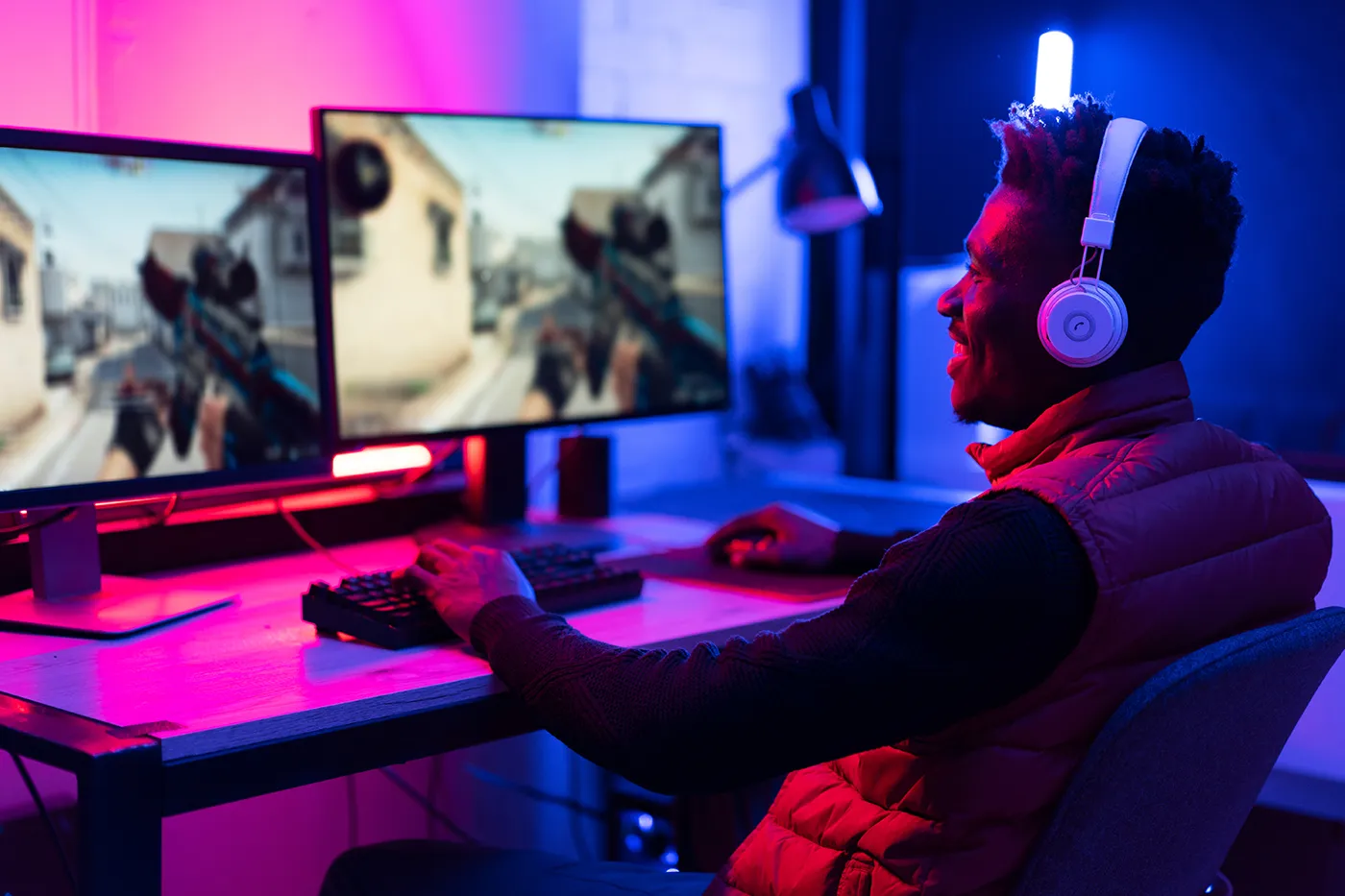 Person gaming at a desk with dual monitors, wearing headphones in a colorful, neon-lit room.