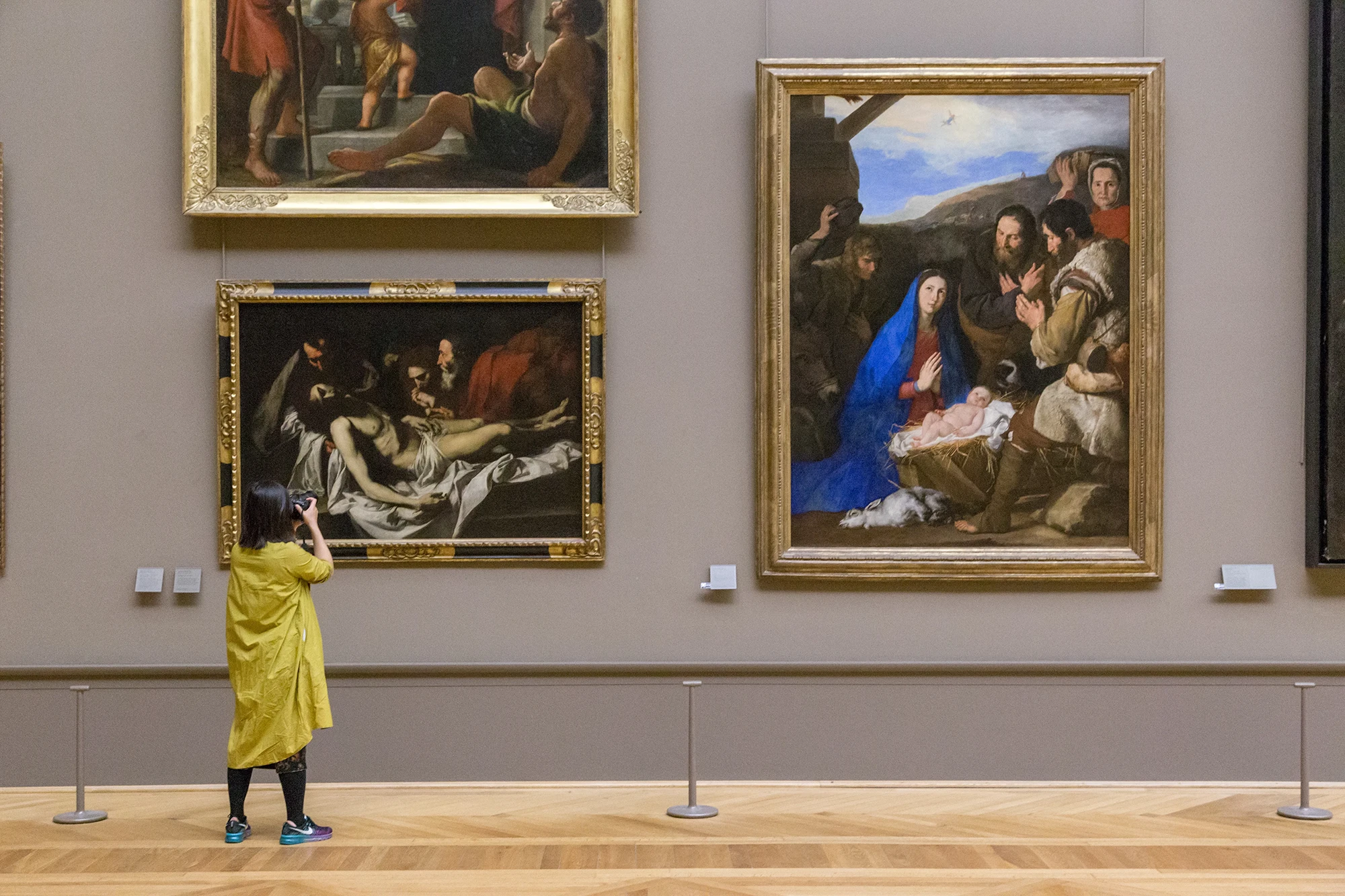 A woman in a yellow coat taking a photo of classical paintings in a museum.