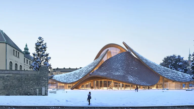 Snow-covered modern curved building with warm glass interior, adjacent historic stone wall and trees.