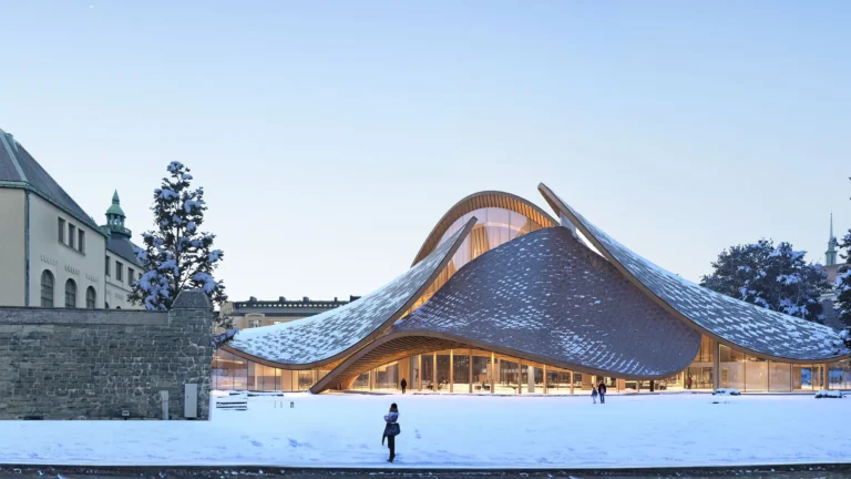 Snowy plaza with sculptural curved building, glass walls, and people walking.