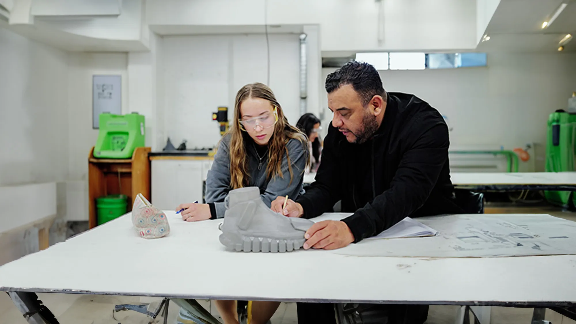 Two people sketching and inspecting a gray shoe form at a white table in a workshop.