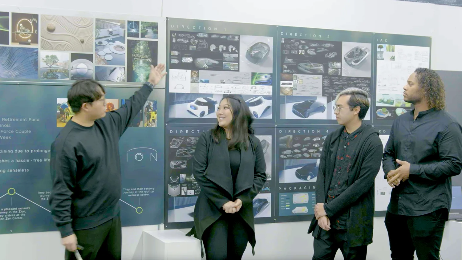 Four Industrial Design Students stand before a wall of design boards; one man points to the boards while others listen and smile.