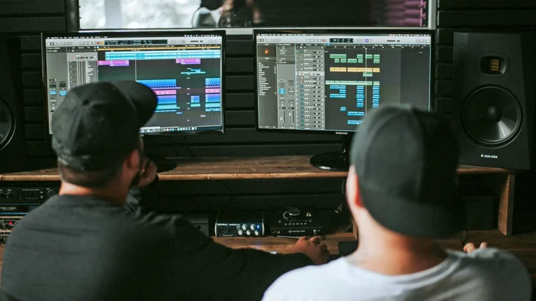 Two ArtU student producers wearing caps work at a dual-monitor audio editing setup with colorful tracks on screen.