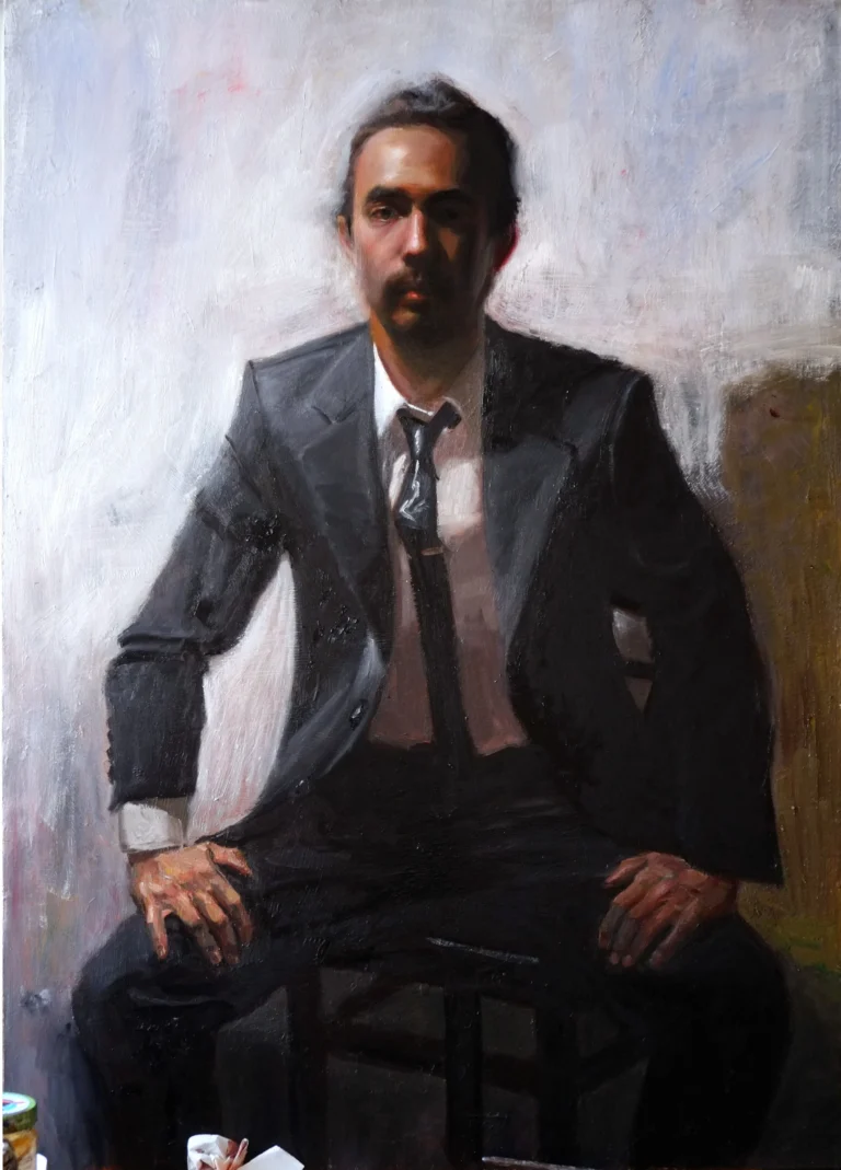 A man in a dark suit sits, hands resting on knees, serious expression