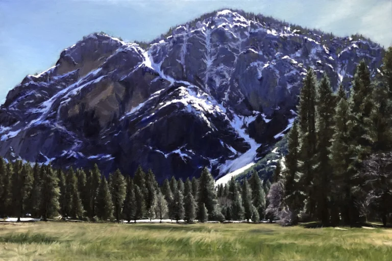 Snow-dusted rocky mountain rises from pine forest under blue sky.