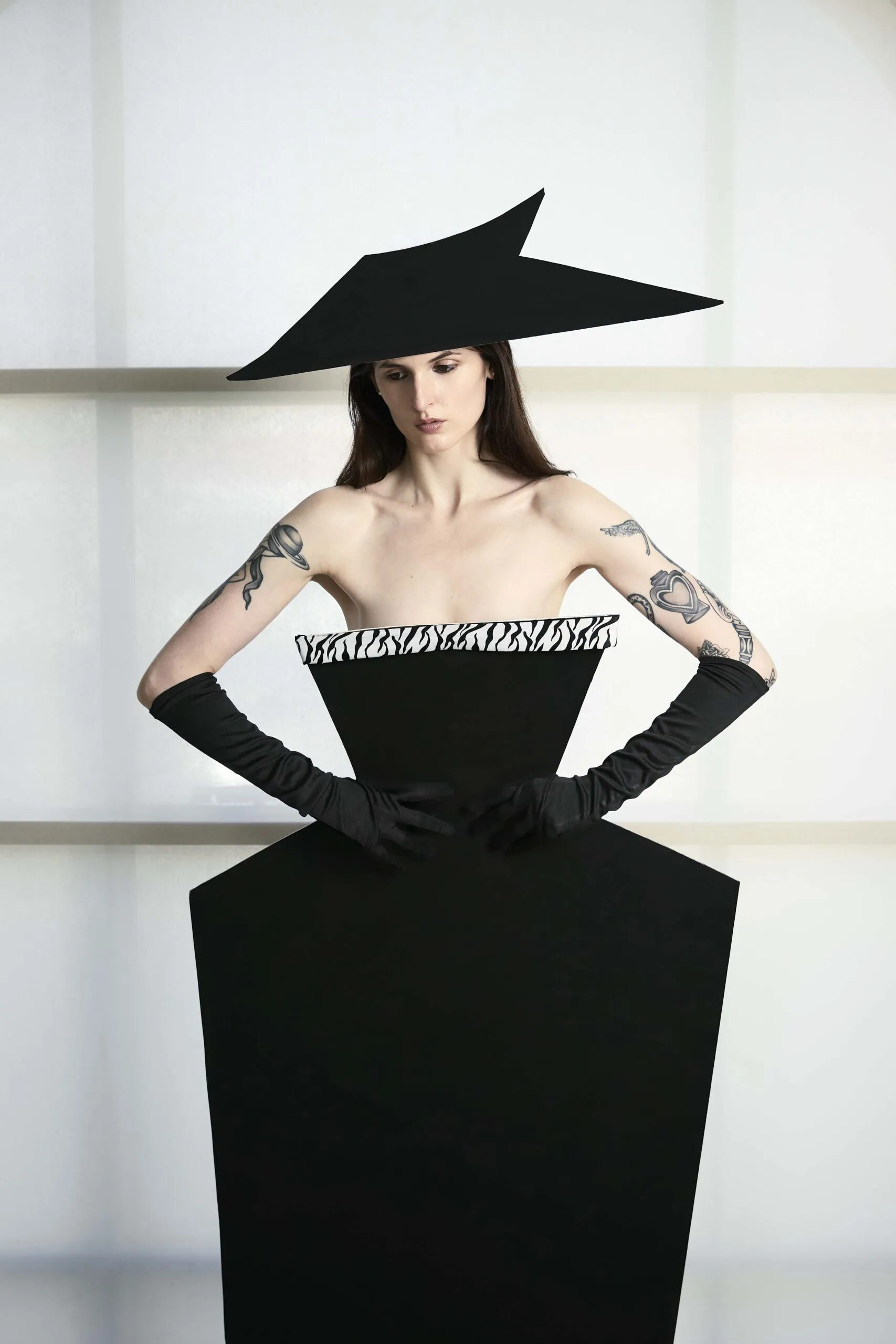 Woman in a black geometric dress and oversized hat, arms akimbo.