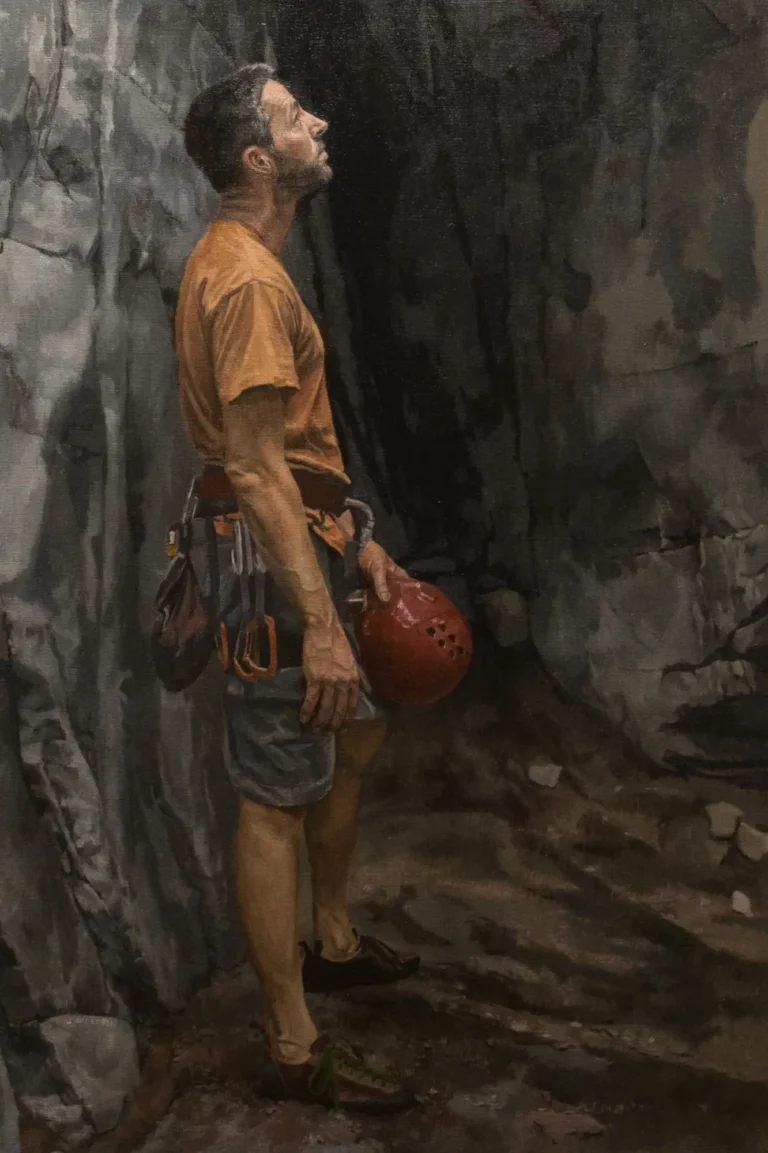 Climber in orange shirt stands, holding red helmet, rests against rock wall
