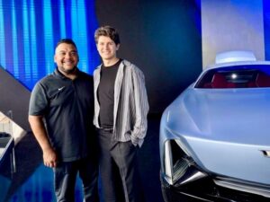 Two men stand smiling beside a futuristic blue car indoors.