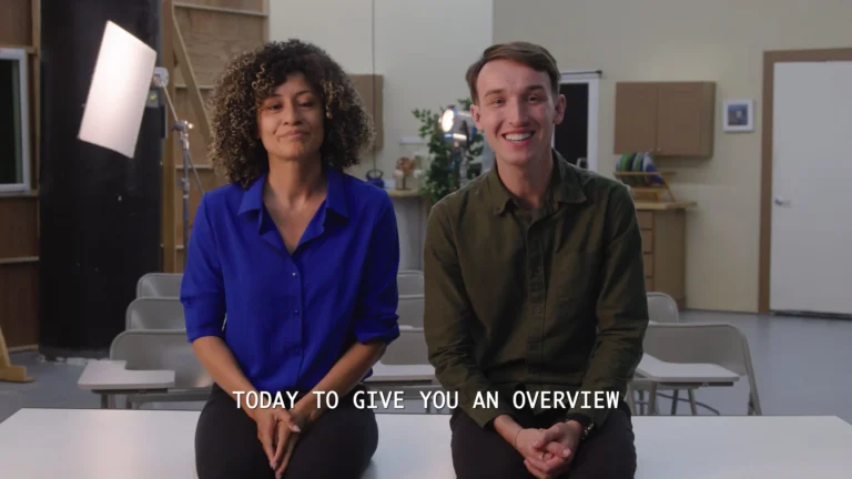 Two smiling people sit side by side in a studio classroom setting.