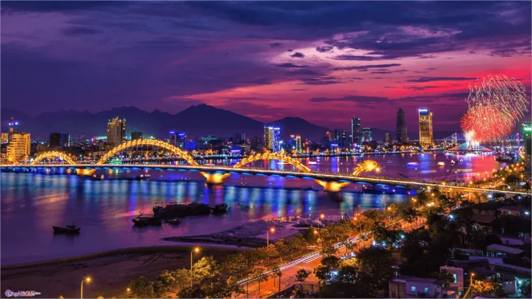 Colorful nighttime cityscape with illuminated bridge, skyline, and fireworks over water.
