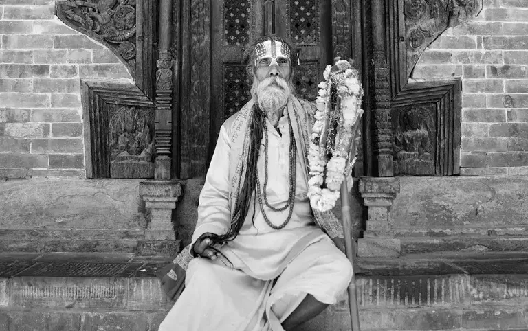 Older man with long beard and vertical forehead markings sits on steps in robes, garlanded staff beside him.