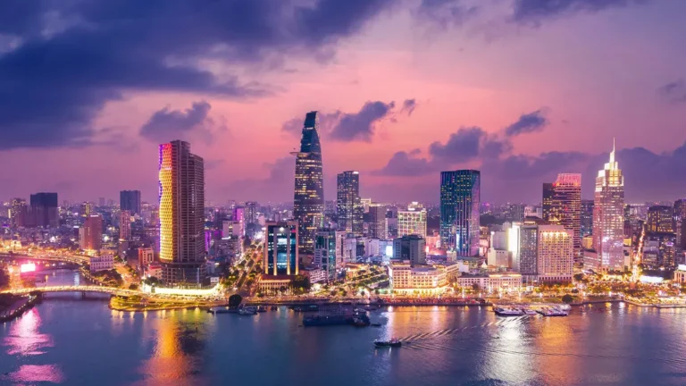 Vibrant city skyline at dusk with illuminated skyscrapers beside a river.