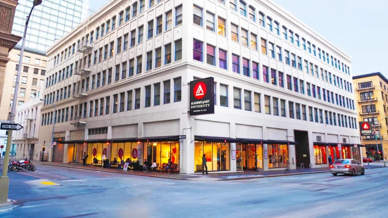 Corner building with large white facade, Academy of Art University sign, ground-floor shops, street with students and cars.