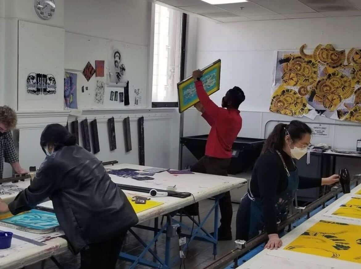 People are engaged in screen printing, holding screens and using tools on tables covered with art materials. The room has art on the walls and a clock shows the time.