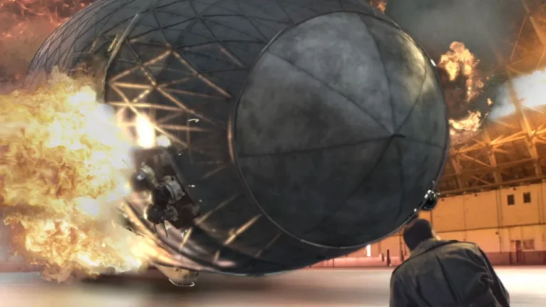 A large, metallic, spherical structure is exploding in an industrial warehouse, surrounded by flames and debris, with a person observing from the foreground.