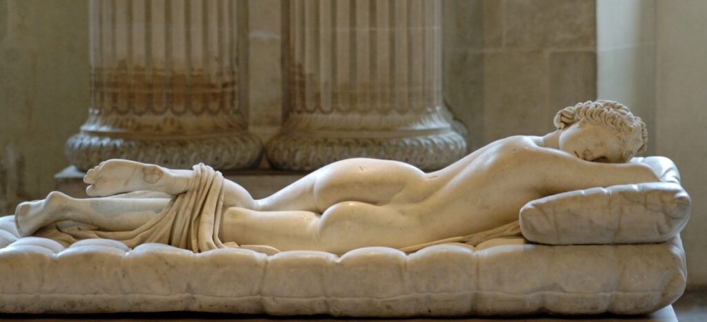 A marble sculpture of a reclining figure on a cushioned surface, resting in a serene pose. The background features ornate columns, suggesting a classical setting. No text is present.