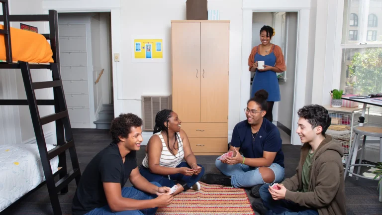 Four people sit on a rug playing cards in a dorm room, while another person stands in the doorway holding a mug, smiling. Bright and tidy room with bunk bed, cabinet, and window.