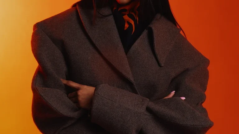 A person stands with arms crossed, wearing a thick, brown coat, ornate red sunglasses, and dark patterned pants against a vibrant orange background.