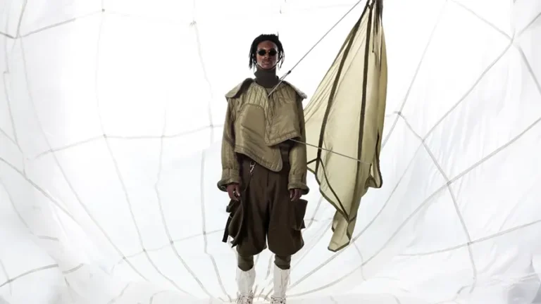 Person wearing layered, earth-toned fashion stands in a bright, curved, net-like structure, holding a draped, translucent fabric. The setting suggests a modern, minimalist artistic theme.