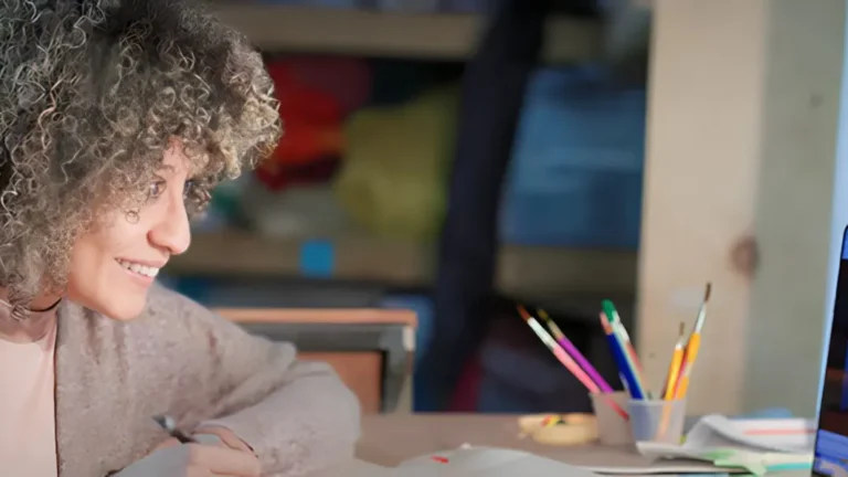 A person smiles while looking at a laptop displaying a bridge image. Nearby, colorful pencils are in a holder on a desk, with shelves and fabric in the background.