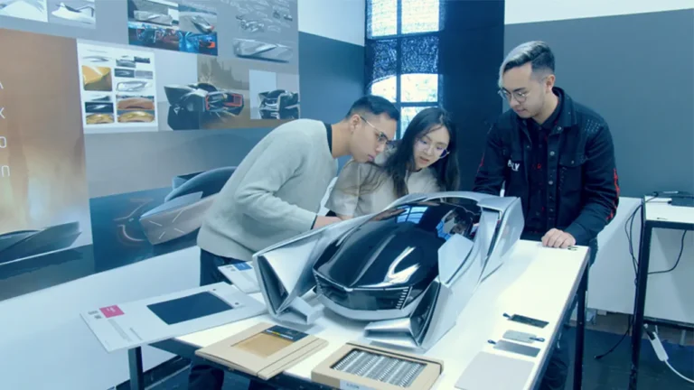 A futuristic car model is examined by three people leaning over a table with design sketches and textures; multiple car concept images adorn the background wall, providing design context.