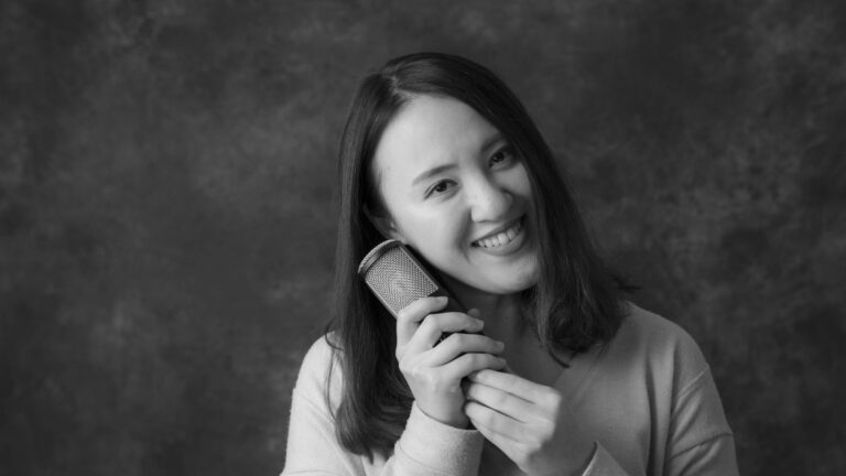 A person holding a microphone smiles warmly, captured in black-and-white against a textured, dark backdrop. The image suggests a joyful mood, possibly related to singing or speaking. No text present.