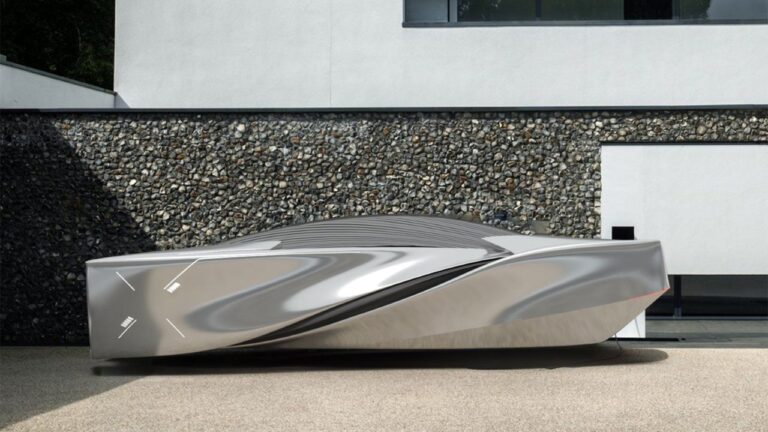 A sleek, futuristic, metallic structure sits on a smooth surface in front of a modern building with stone-textured lower walls and plain upper walls. There is no text in the image.