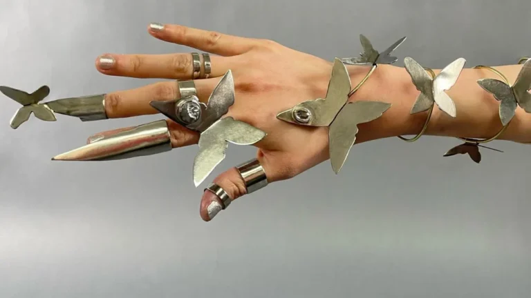 A hand wears metallic rings and finger extensions shaped like butterflies, resting against a neutral gray background. Rings are adorned with intricate butterfly designs, creating an artistic and whimsical appearance.