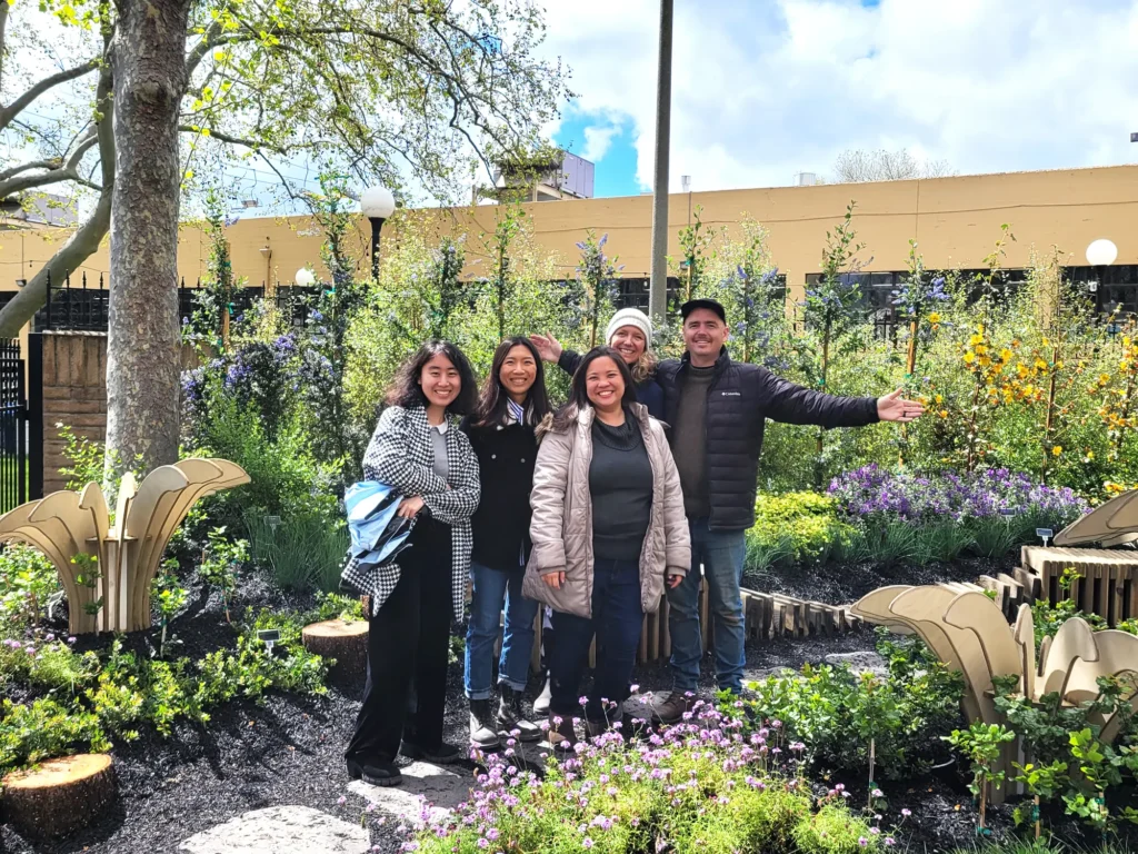 Group of five people smiles and poses in a vibrant garden, surrounded by lush greenery and colorful flowers, with a yellow building and cloudy sky in the background.