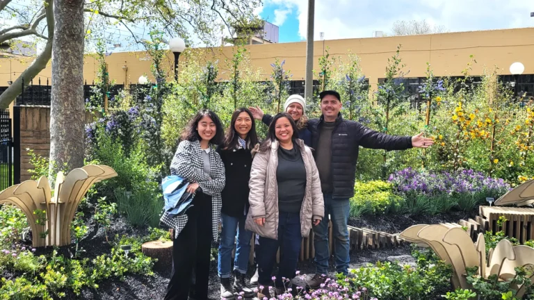Group of five people smiles and poses in a vibrant garden, surrounded by lush greenery and colorful flowers, with a yellow building and cloudy sky in the background.