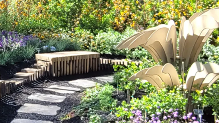 A wooden bench sits stationary beside a stone path in a vibrant garden filled with lush greenery and colorful flowers under a clear sky.