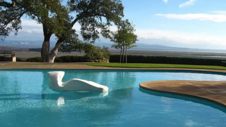 A white sculptural chair floats in a clear blue swimming pool, surrounded by a patio and lush greenery, with an expansive view of distant hills under a partly cloudy sky.