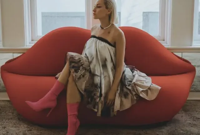 A person sits gracefully on a red, lip-shaped sofa, wearing a strapless dress and pink boots, in a room with large windows.