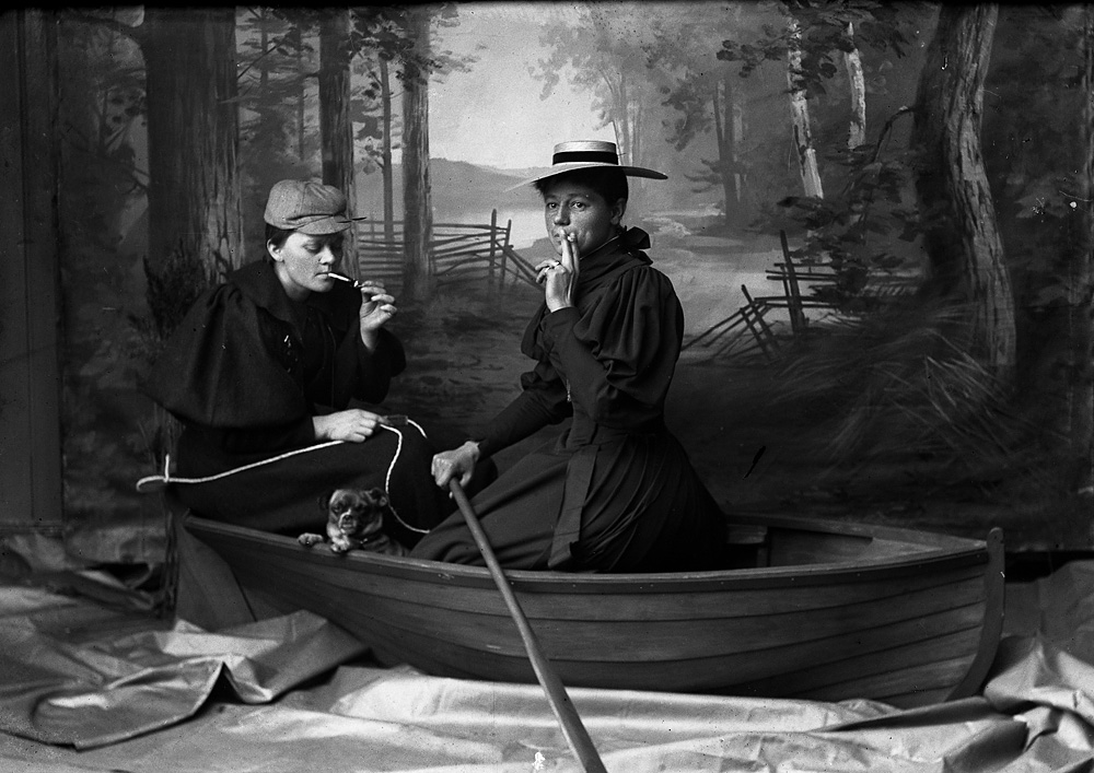 Two people in 19th-century attire, smoking pipes, sit in a small boat with a puppy. A painted backdrop depicts a forest and fence, creating an outdoor illusion.