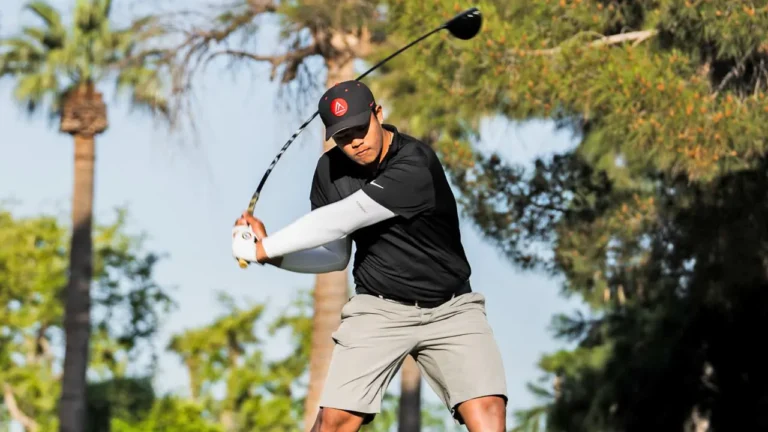 Golfer swings a club in mid-motion on a sunny day, surrounded by tall palm trees and greenery. The golfer wears a black shirt, gray shorts, and a cap with a red logo.