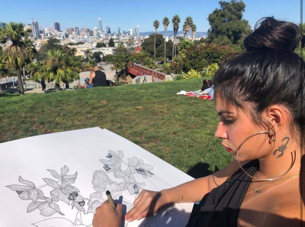 Artist sketches flowers on paper, sitting on grass in a park with palm trees and city skyline in the background.