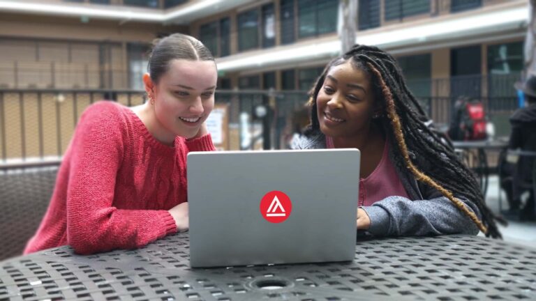 Two people are smiling and looking at a laptop displaying a red and white logo, seated outside at a metal table, with a blurred building in the background.
