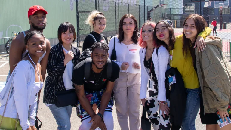 A group of nine people poses smiling, with some wearing name tags. They stand outdoors near a basketball court, under sunny weather, next to a green wall.