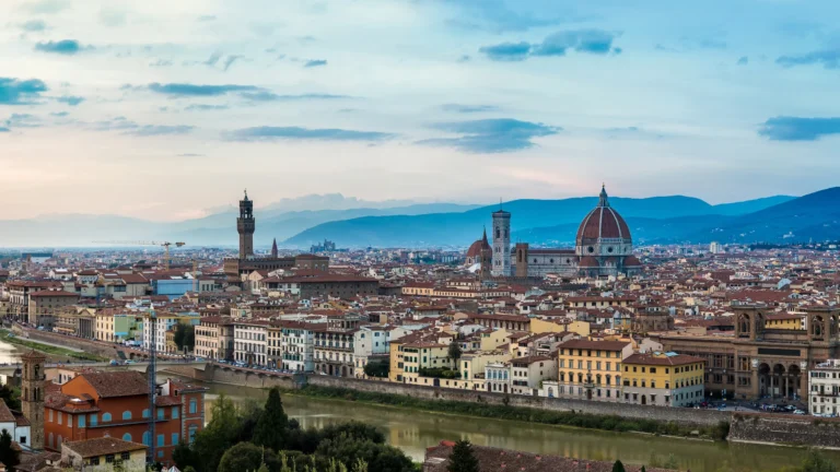 A cityscape features Florence's iconic red-domed cathedral and medieval towers. Buildings line the river beneath a vast, partly cloudy blue sky, with distant mountains in the background. No text present.