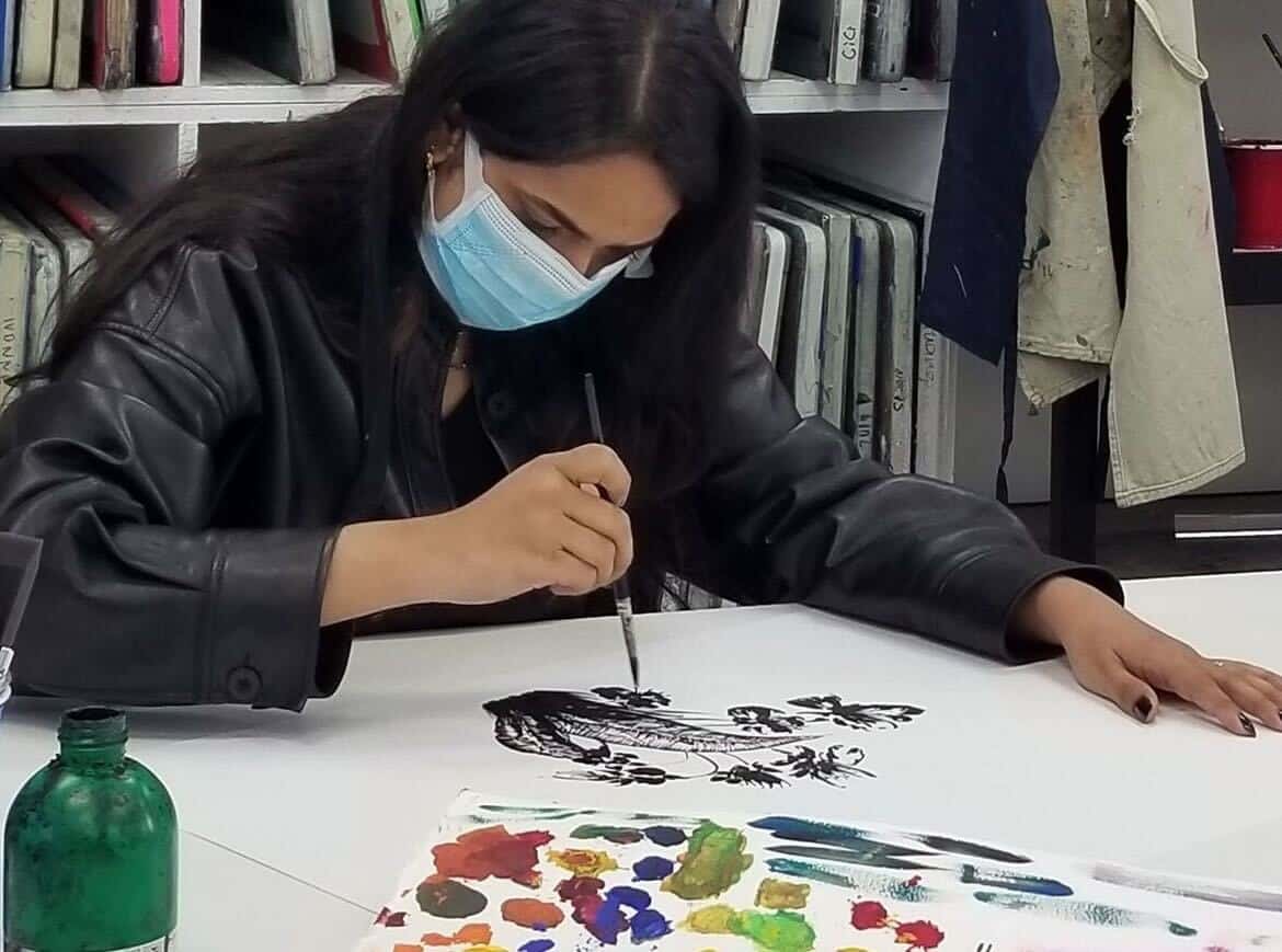 Artist wearing a mask paints with a brush, creating a detailed black-and-white design on paper. Colorful palette and green bottle visible. Shelves with books and hanging cloth in the background.