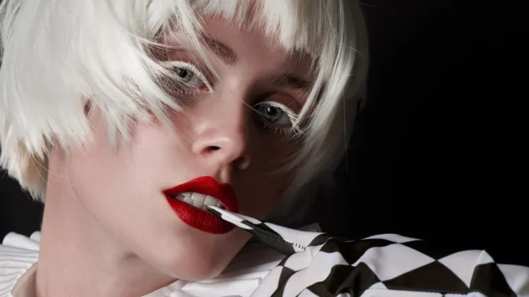 Person with short white hair and red lips holds a dagger in their mouth. They wear a ruffled white collar and checkered, glove-covered arm against a dark background.