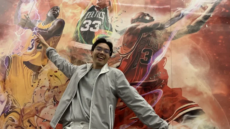 A person poses energetically, arms extended, in front of a dynamic mural featuring basketball players in vibrant action. Players wear jerseys with numbers 32, 33, and 23, amidst swirling colors.