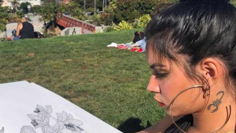 A person with large hoop earrings and a scorpion tattoo draws flowers on a sketchpad in a sunny park, with a city skyline and palm trees in the background.
