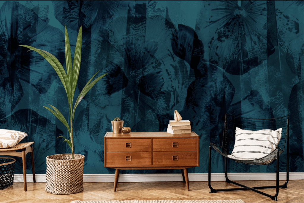 A mid-century modern wooden dresser stands in a room, adorned with stacked books and plants, beside a wicker basket and a black wire chair with a striped pillow. The background features a dark teal floral-patterned wall.
