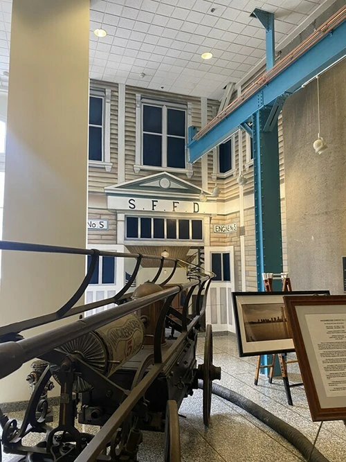 Historic fire engine displayed indoors with a façade reading "S.F.F.D," representing an old fire station. Nearby informational placards stand on easels in a room with high ceilings and metal beams.