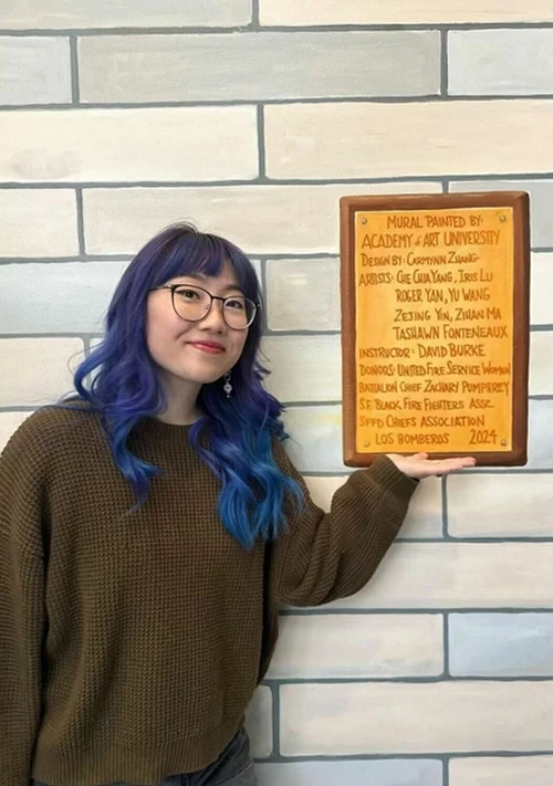 A person with blue hair smiles and points to a yellow plaque on a brick wall. The plaque reads: 

"MURAL PAINTED BY: ACADEMY OF ART UNIVERSITY  
Design by: Guanxun Zhang  
Artists: Ge Guo, Yang, Iris Lu,  
Roger Yan, Yu Wang,  
Zeying Yin, Zixuan Ma  
Tasiann Fontenaux  
Instructor: Davi Burke  
Donors: United Fire Service Women, Battalion Chief Zachary Mummey,  
SFBlk FFs, Fire Fighters Assc.,  
SPFD Chiefs Association, Los Bomberos  
2024"