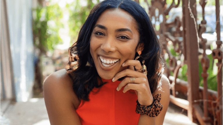 A person smiles brightly, seated and leaning on their hand outdoors, surrounded by lush greenery and decorative metalwork, wearing a vibrant red outfit and multiple rings.
