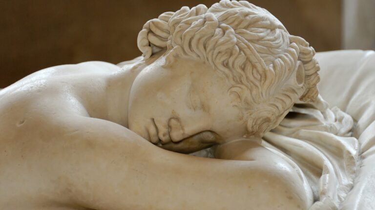 Marble sculpture of a sleeping figure with curled hair rests head on folded arms. Draped fabric details the context, suggesting serenity in a classical setting. No text is present.